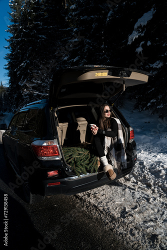 A young woman in sunglasses poses in the trunk of a car with a pine bouquet. Concept for a winter trip to the mountains