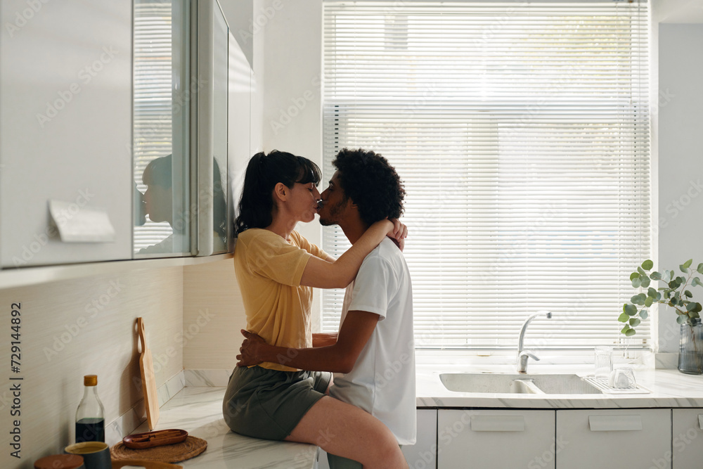 Young affectionate woman in casualwear sitting on kitchen counter and kissing her husband keeping his hands around her waist Young affectionate woman in casualwear sitting on kitchen counter and kissing her husband keeping his hands around her waist