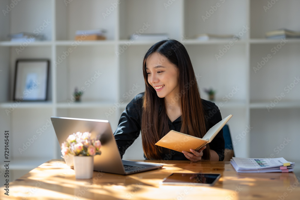 Attentive businesswoman jotting down notes while working on a project with her laptop at a sunny table.. Attentive businesswoman jotting down notes while working on a project with her laptop at a sunny table..