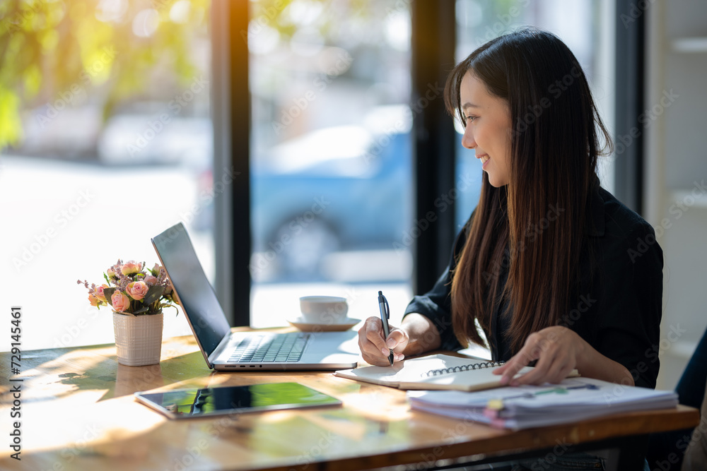 Obraz premium Attentive businesswoman jotting down notes while working on a project with her laptop at a sunny table..