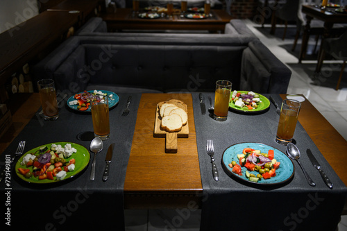 Greek salad. A set table with cutlery for four people in a restaurant. Fresh greek salad with tomato, cucumber, bell pepper, olives, and feta cheese on blue plate, top view, dark table.