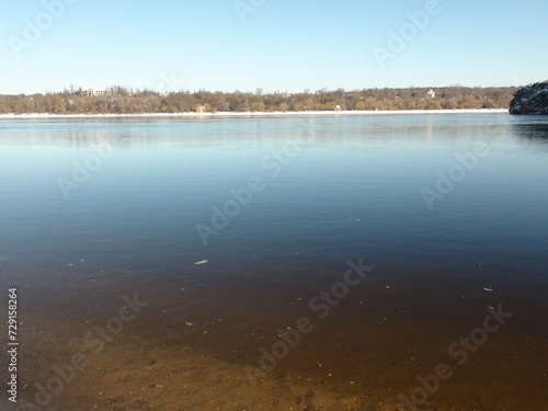 A panorama of the transparent water surface of the wide Dnieper River  through which a clean sandy bottom surrounded by a snow-covered shore shines through.