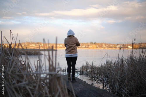 A girl in a hood and a warm jacket, standing on a wooden footbridge with a phone in the middle of reeds near the lake, in the cold season