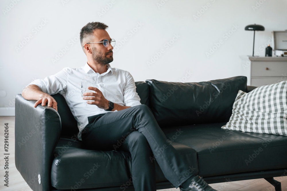 Sitting on the sofa. Elegant man in business clothes is indoors with glass of water Sitting on the sofa. Elegant man in business clothes is indoors with glass of water