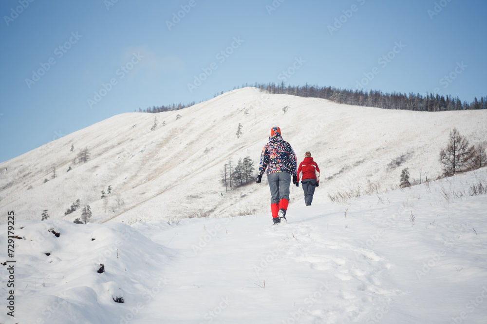 Two people walking up a snowy hill on a bright winter day with snow-covered trees