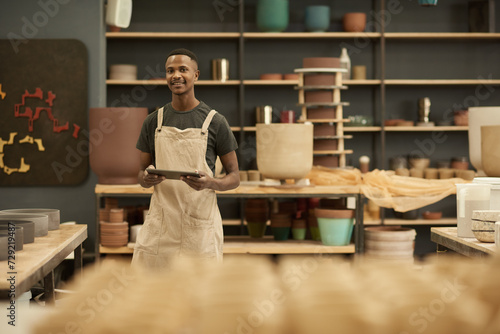 Young African potter with a tablet smiling in a large ceramics workshop