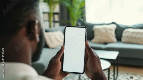 An African American man is seen in a living room setting, holding a phone with a fully white screen in a shot taken from over the shoulder photo