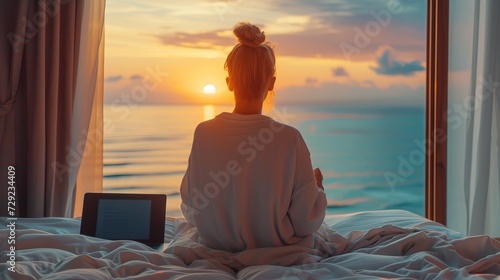 A tourist woman sitting on bed, looking to Beautiful sunset above sea or ocean, reflection of sun in the water after video conference meeting with colleagues.