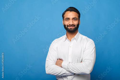 A poised Indian businessman in a crisp white shirt stands with crossed arms against a blue background, exuding a professional and approachable demeanor. Business, finance, or corporate concept
