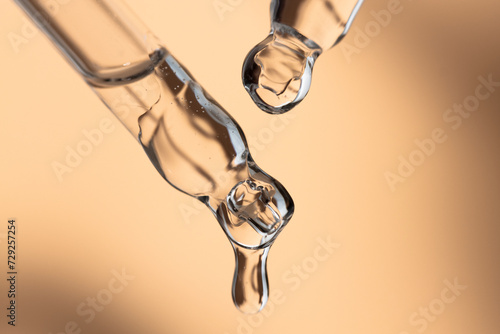 Two glass pipettes with transparent essential oil dripping on a beige background, beauty background.