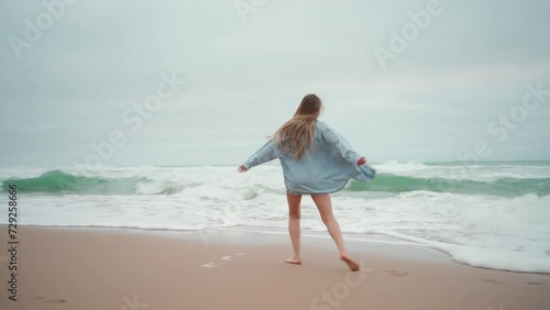 Full length of carefree woman with blond hair running and turning along sea on sandy beach. Female tourist in denim shirt on summer vacation. Excited lady is on beach holiday.