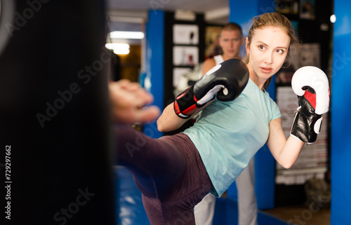 Woman in boxing gloves kicking punchbag during training. Trainer standing behind and watching.