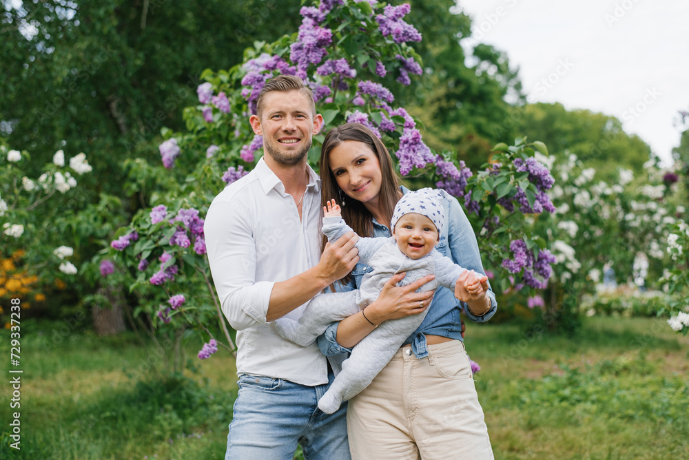 Obraz premium Mother, father and little son are walking in a spring blooming park. A happy mom and dad with a small child are having fun near the lilac. The concept of a happy family.