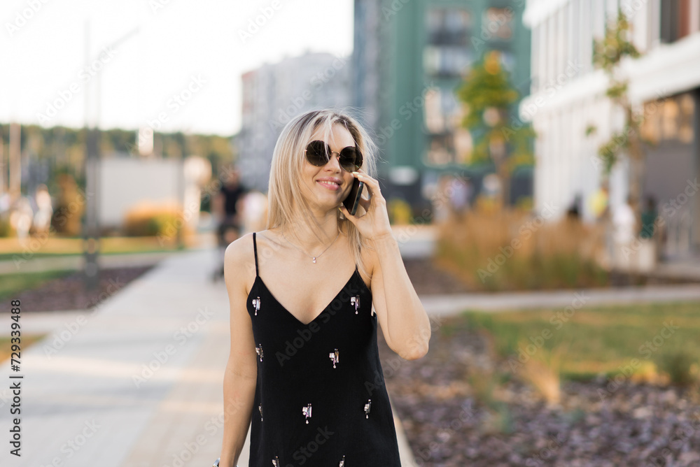 Young woman in a black dress, talking on a cell phone, walking on a city street in the summertime