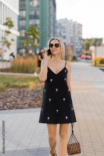 Young woman in a black dress, talking on a cell phone, walking on a city street in the summertime