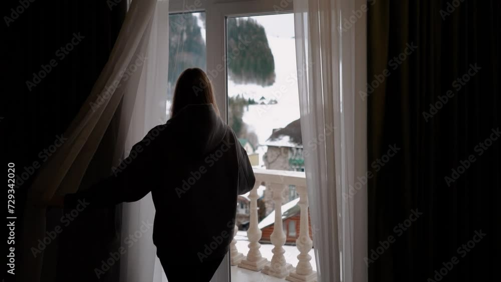 a girl near the window opens the curtain in a hotel room overlooking the mountains. Ski resort with chalets, spa and relaxation. Vacation and holidays in winter in the forest a girl near the window opens the curtain in a hotel room overlooking the mountains. Ski resort with chalets, spa and relaxation. Vacation and holidays in winter in the forest