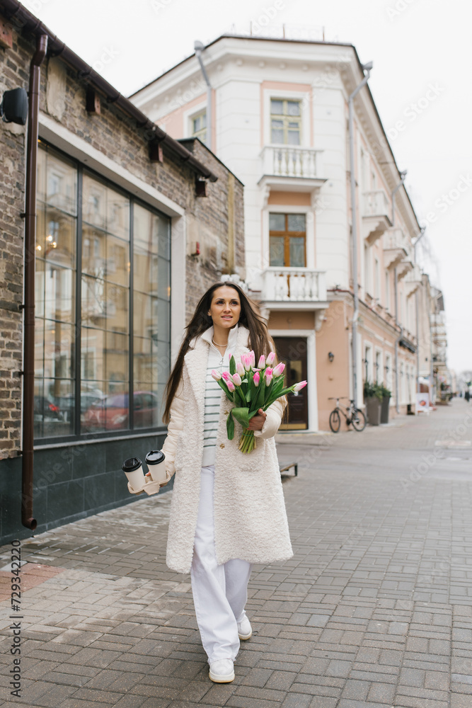 Young stylish woman in a light-colored coat walks down a city street with a paper cup of coffee or tea and a bouquet of spring tulips