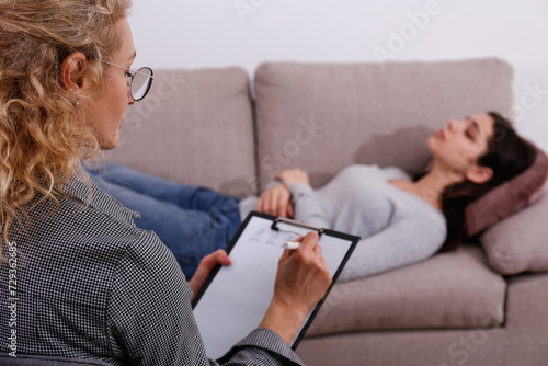 Therapy session concept. Adult female therapist taking notes in a notebook while her client is sharing her thoughts. Close up, copy space, background.