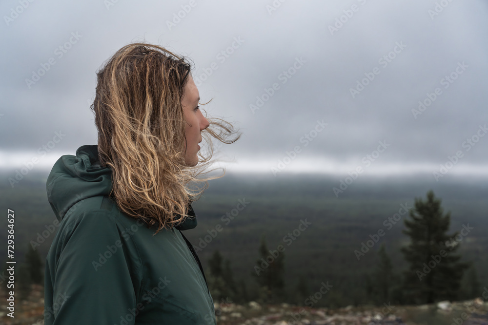 Side face view of Caucasian woman with blond hair blowing in wind in nordic nature, female hiker standing on mountain, in mind looking away over the forest and the stormy clouds in the background Side face view of Caucasian woman with blond hair blowing in wind in nordic nature, female hiker standing on mountain, in mind looking away over the forest and the stormy clouds in the background