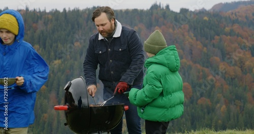 Adult man takes meat out of the barbecue grill for children. Multiethnic group of travelers or big family resting outdoors during vacation trip. Mountains landscapes and forest in the background.