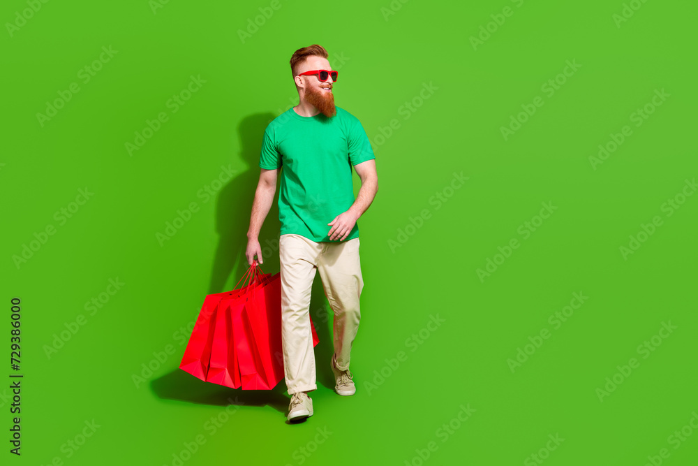 Full length portrait of cheerful young man hold mall bags walk empty space ad isolated on green color background Full length portrait of cheerful young man hold mall bags walk empty space ad isolated on green color background