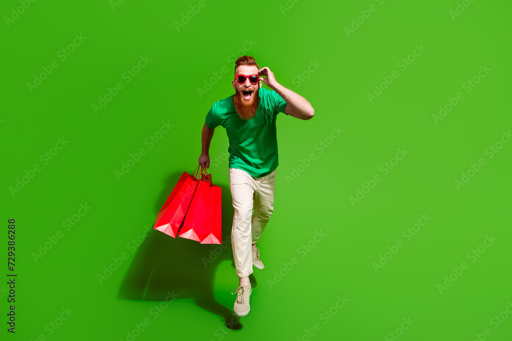 Full length portrait of handsome impressed guy hold store bags jump run empty space isolated on green color background Full length portrait of handsome impressed guy hold store bags jump run empty space isolated on green color background