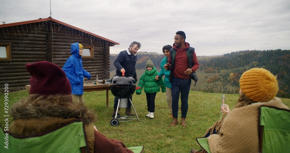 Diverse family together cook tasty dinner on BBQ grill on beautiful hill. Group of outdoor enthusiasts resting outdoors during vacation trip in mountains. Concept of outdoor exploration and tourism.