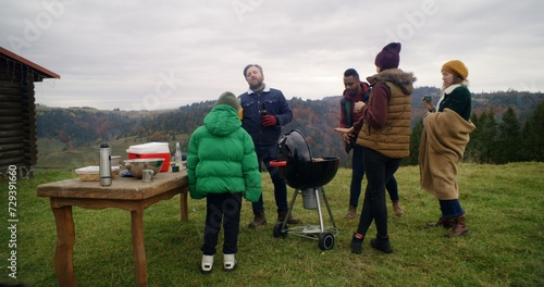 Multicultural family stand together near BBQ grill and discuss trip on beautiful hill. Group of hiking buddies stopped to rest after long expedition at mountains. Active leisure and tourism concept.