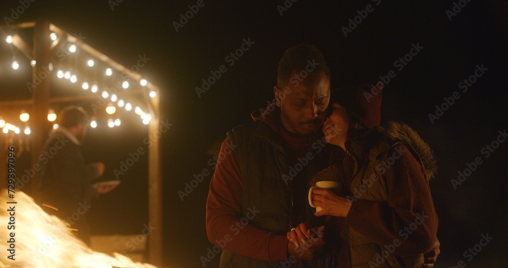 Multiethnic couple drinks tea, kisses and hugs while standing near the fire outdoors during vacation trip. In the background traveling friends rest in stylish wooden gazebo decorated with light bulbs. Multiethnic couple drinks tea, kisses and hugs while standing near the fire outdoors during vacation trip. In the background traveling friends rest in stylish wooden gazebo decorated with light bulbs.