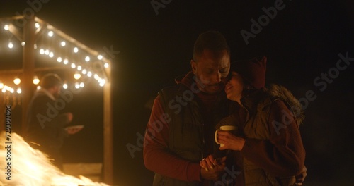 Multiethnic couple drinks tea, kisses and hugs while standing near the fire outdoors during vacation trip. In the background traveling friends rest in stylish wooden gazebo decorated with light bulbs.