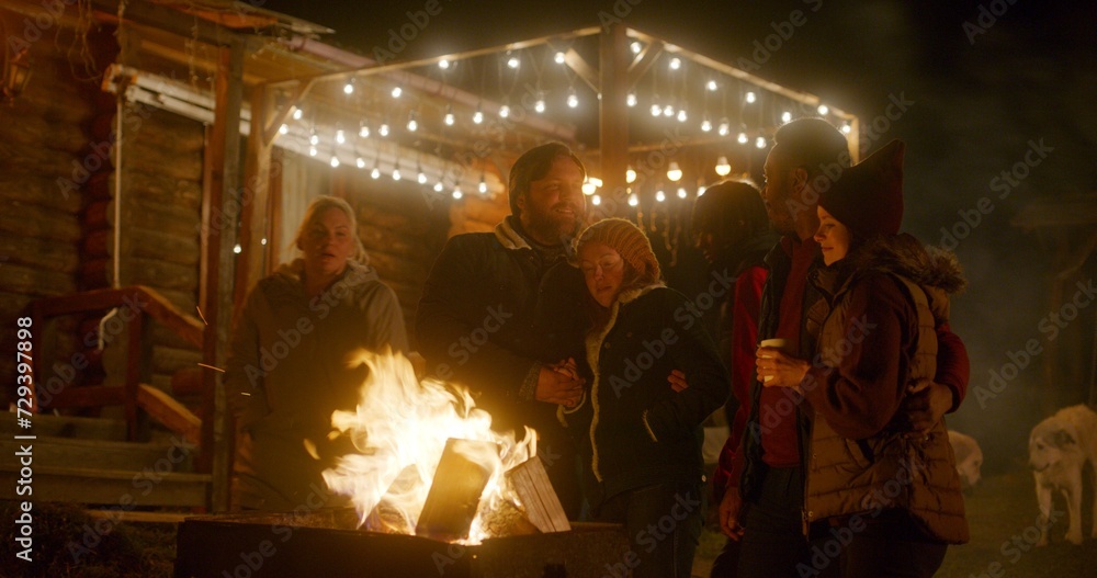 Multiethnic tourist couples stand near the fire outdoors in the evening during vacation. Friends or family of travelers resting together outside of stylish wooden house decorated with light bulbs.