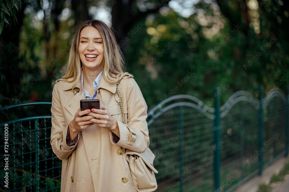 Woman laughing while using phone and walking on a city street near a park.