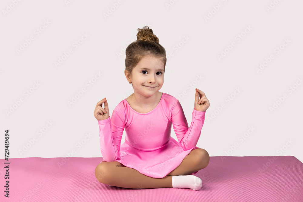 Little girl gymnast in lotus position smiling beautifully to camera meditating, sitting on pink fitness mat wearing pink gymnastic dress.
