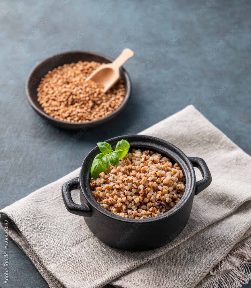 Buckwheat porridge with fresh herbs in a black pan on a napkin on a dark background with raw grains. Buckwheat porridge with fresh herbs in a black pan on a napkin on a dark background with raw grains.