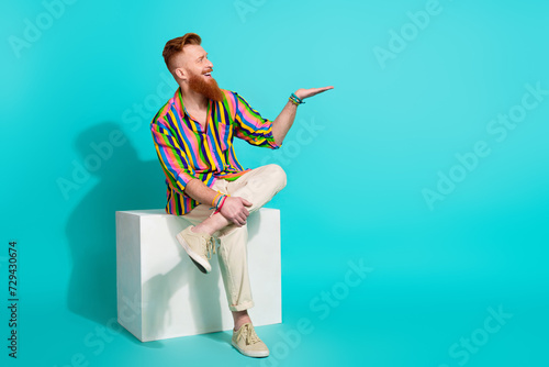 Full body length photo of red hair macho hipster sit podium holding arm new perfume advertisement isolated on cyan color background