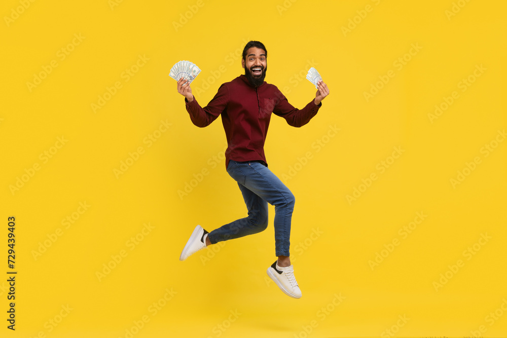 Excited millennial indian guy holding money cash in hands, jumping