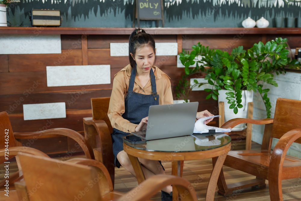 Woman, cute Asian woman, businessman, small cafe owner, beige shirt sitting in front of his notebook and checking the sales documents of the cafe thoughtful expression. It's inside minimalist cafe. Woman, cute Asian woman, businessman, small cafe owner, beige shirt sitting in front of his notebook and checking the sales documents of the cafe thoughtful expression. It's inside minimalist cafe.