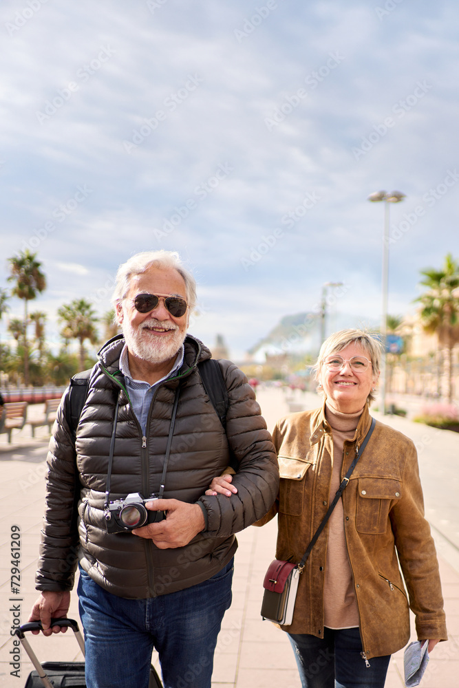 Vertical smiling Caucasian mature tourist couple standing posing looking at camera in street with their luggage. Husband and wife enjoying their retirees holidays wearing winter clothes on sunny day Vertical smiling Caucasian mature tourist couple standing posing looking at camera in street with their luggage. Husband and wife enjoying their retirees holidays wearing winter clothes on sunny day