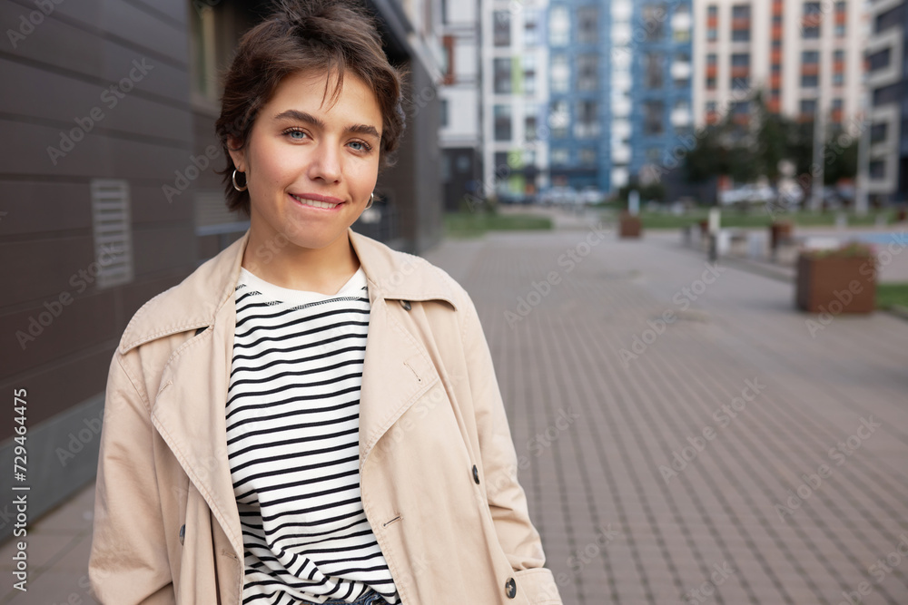 Cute stylish girl city tourist in trench standing on street in residential area, walking alone, looking at camera biting her lower lip like having crazy adventurous idea. Copy space on the left Cute stylish girl city tourist in trench standing on street in residential area, walking alone, looking at camera biting her lower lip like having crazy adventurous idea. Copy space on the left