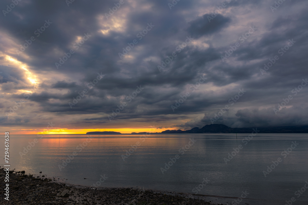 Bright orange sunrise on the coast with dark dramatic clouds billowing over mountains in the distance. Beaumaris, North Wales