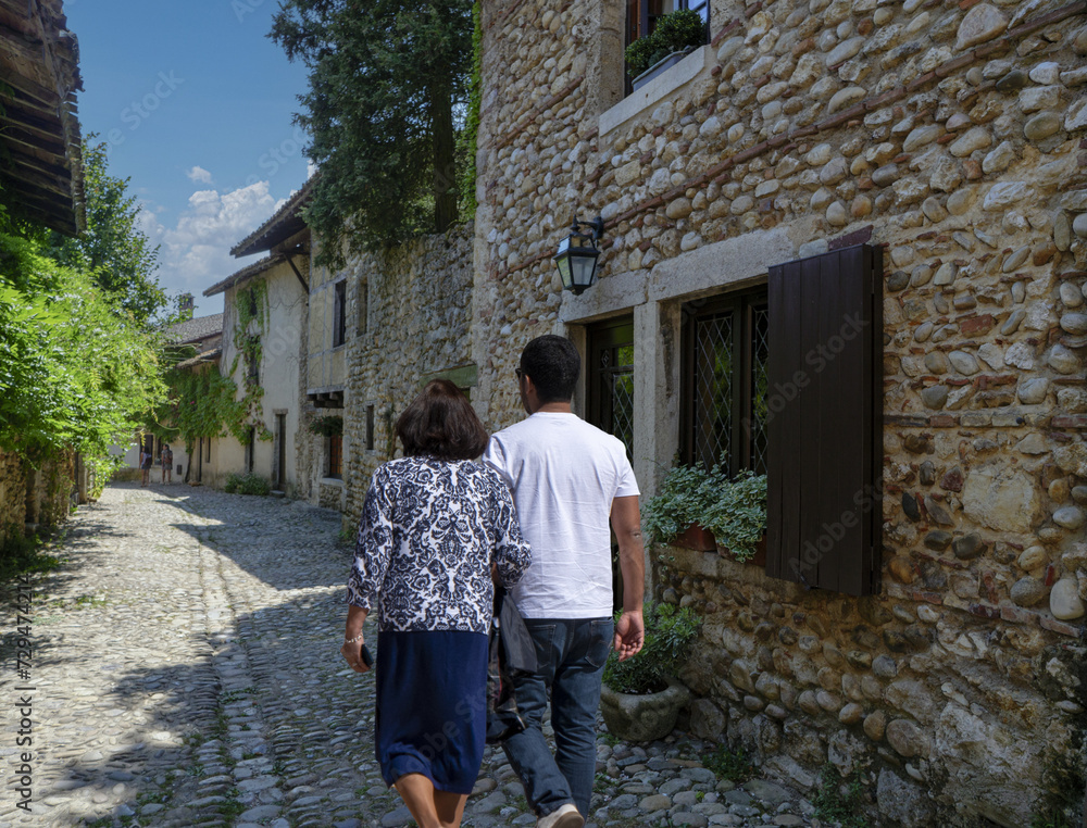 Couple qui se promène en été dans une ruelle pavée d'un village médiéval, la rue est bordée de murs en pierres avec des fleurs aux fenêtres 