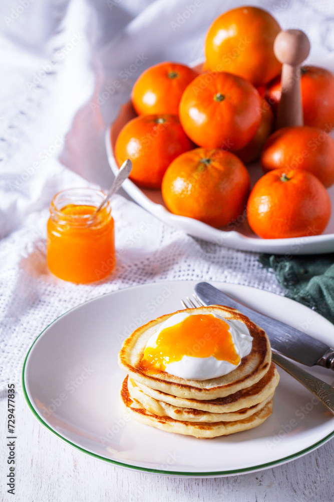 Pancakes served with tangerine jam and sour cream, and tangerines on a light background.
