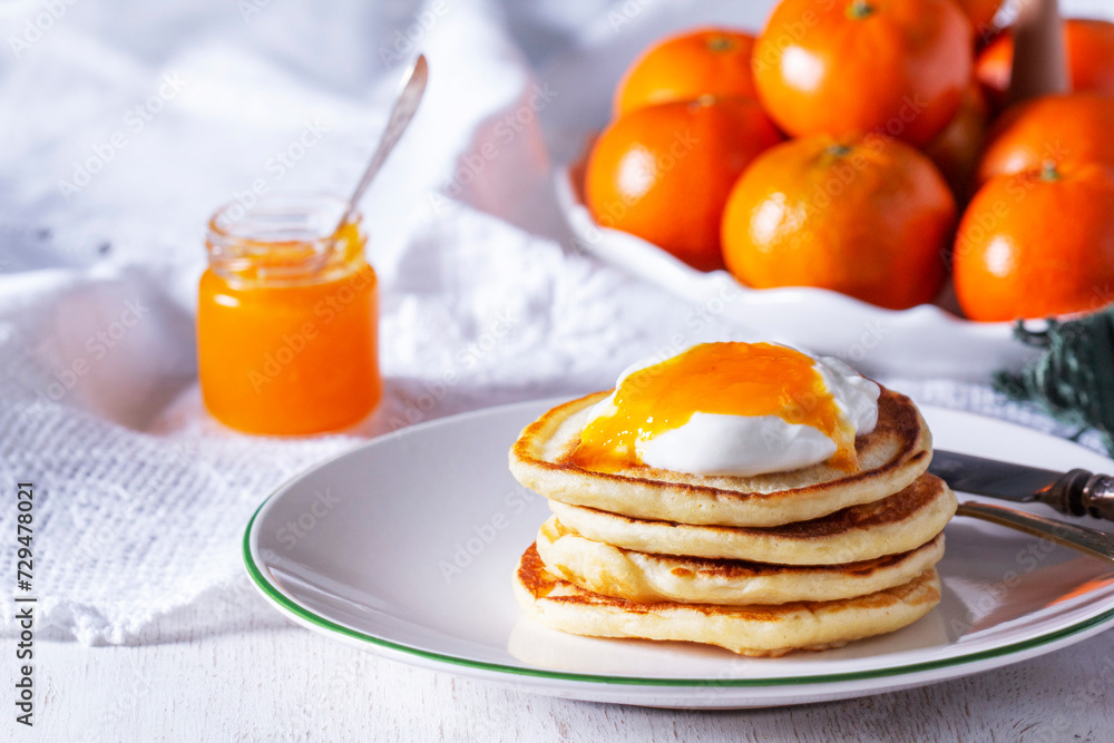 Pancakes served with tangerine jam and sour cream, and tangerines on a light background.