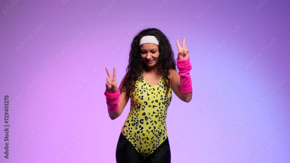 Nerdy curly woman in a bright colored outfit from the 80s shows a victory gesture and dances with a smile on a purple background. Symbolizing victory and joy. Nerdy curly woman in a bright colored outfit from the 80s shows a victory gesture and dances with a smile on a purple background. Symbolizing victory and joy.