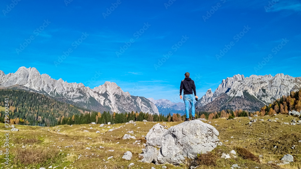 Man standing on rock formation on golden alpine meadow in autumn. Scenic view of majestic mountains of Carnic Alps in Sauris di Sopra, Friuli Venezia Giulia, Italy. Tranquil atmosphere in Italian Alps