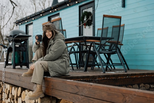 Countryside winter vacation for the weekend. Woman with a mug of warming drink against the background of a rented wooden house with a gas grill and Christmas decorations