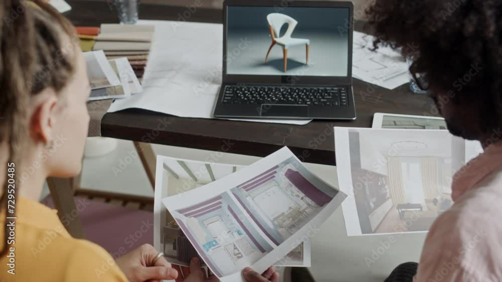 Over shoulder shot of two ethnically diverse interior designers having discussion while looking at 3d rendering pictures of modern apartment design project, co-working in office at daytime