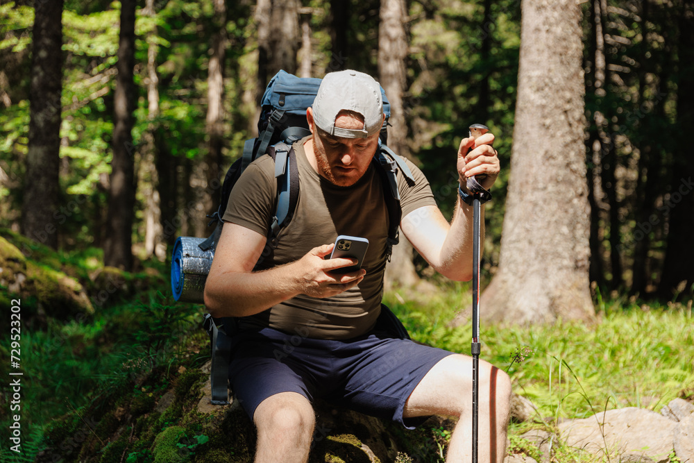 Adult man with smartphone backpack trekking stick sitting in green forest