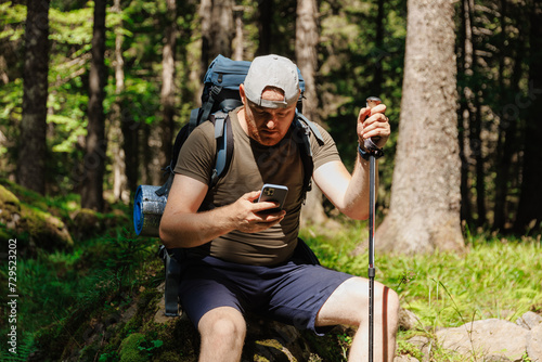 Adult man with smartphone backpack trekking stick sitting in green forest