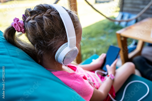 Toddler girl sitting in cafe with headphones and mobile phone in her hands playing games